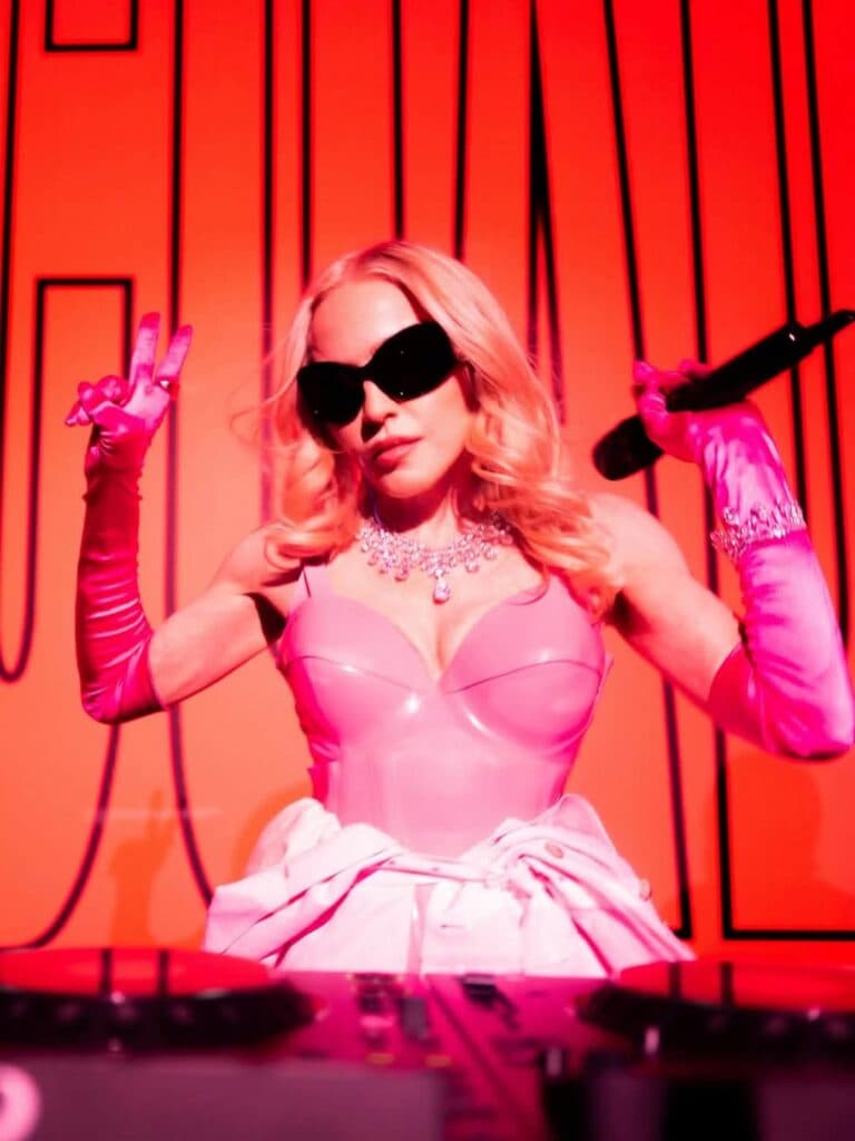 Madonna en un escenario con fondo rojo, luciendo un atuendo rosa y gafas oscuras, sosteniendo un micrófono y haciendo una señal de paz.